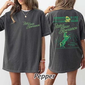 May include: Dark grey t-shirt with the text "Star Lasso Experience" in green on the front. The back of the shirt features the text "Jupiter's Claim" and a green graphic of a rearing horse. The shirt also has the text "Friday September 9th at 8PM".