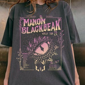 May include: Dark grey t-shirt with a pink and yellow graphic design. The design features the text "MANON BLACKBEAK WORLD TOUR" and a stylized eye. The shirt also lists performers' names and the words "No 1 HIT" in the design.