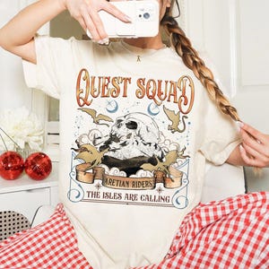Peut inclure: T-shirt cr&egrave;me avec l'inscription "QUEST SQUAD" en lettres orange et marron. Le graphisme repr&eacute;sente un cr&acirc;ne, des chauves-souris et les textes "ARETIAN RIDERS" et "THE ISLES ARE CALLING". La personne porte un pantalon &agrave; carreaux rouges et blancs.