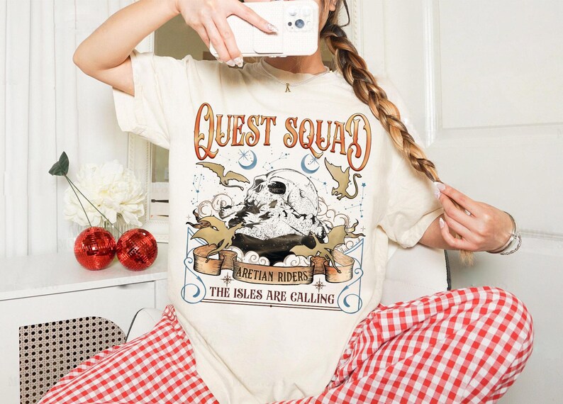 Peut inclure: T-shirt cr&egrave;me avec l'inscription "QUEST SQUAD" en lettres orange et marron. Le graphisme repr&eacute;sente un cr&acirc;ne, des chauves-souris et les textes "ARETIAN RIDERS" et "THE ISLES ARE CALLING". La personne porte un pantalon &agrave; carreaux rouges et blancs.