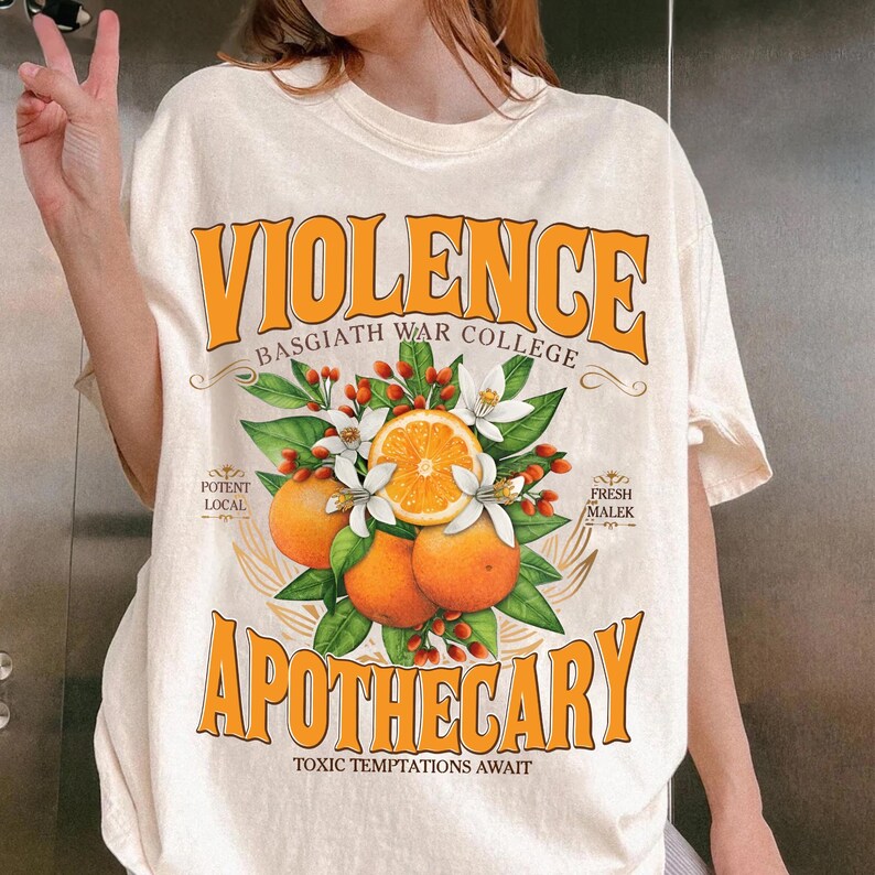 Puede incluir: Camiseta color crema con un dise&ntilde;o gr&aacute;fico naranja. El dise&ntilde;o presenta la palabra "VIOLENCE" encima de "BASGIATH WAR COLLEGE" en texto naranja. Debajo, "APOTHECARY" con una ilustraci&oacute;n naranja y blanca de naranjas y flores.