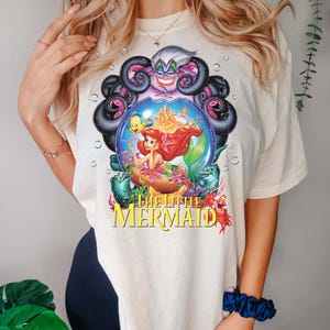 May include: Cream-colored t-shirt featuring a colorful graphic from "The Little Mermaid." The design includes Ariel, Flounder, and Ursula, with the movie title in yellow. The graphic is set within a bubble-like frame.