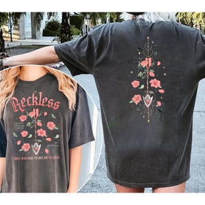 May include: Dark gray t-shirt with the word "Reckless" in a script font, featuring a floral design with pink flowers and the text "Souls who dare to love and be loved."