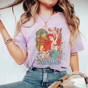 May include: A lilac-colored t-shirt featuring a graphic from "The Little Mermaid." The design includes Ariel, Flounder, and other sea creatures. The words "The Little Mermaid" are printed in a teal color below the illustration. The shirt is paired with blue jeans.