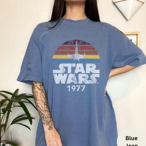 May include: A blue jean t-shirt with a vintage Star Wars graphic. The graphic features a retro sunset with a silhouette of an X-Wing fighter and the text "Star Wars 1977".