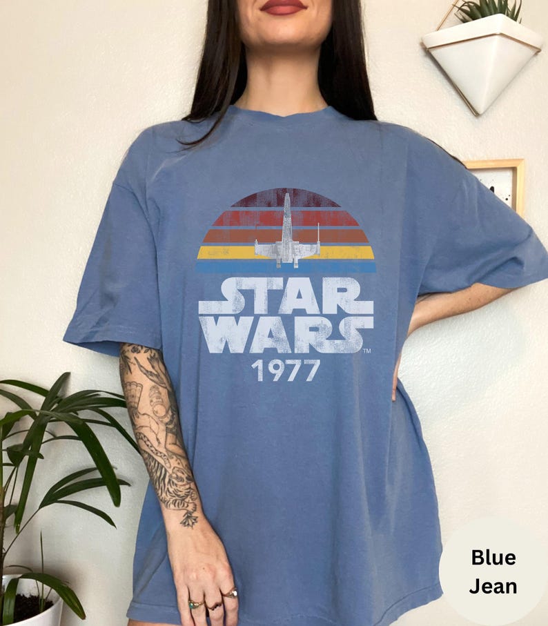 May include: A blue jean t-shirt with a vintage Star Wars graphic. The graphic features a retro sunset with a silhouette of an X-Wing fighter and the text "Star Wars 1977".