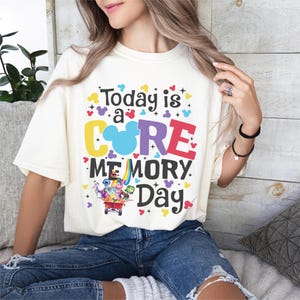 May include: Cream-coloured t-shirt with the text "Today is a CORE MEMORY Day." The design features a Mickey Mouse head, colourful hearts, and characters from the movie Inside Out in a wagon. The shirt is paired with blue jeans.