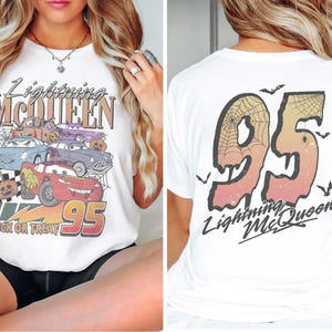 K&ouml;nnte beinhalten: Wei&szlig;es T-Shirt mit einer Grafik von Lightning McQueen und anderen Cars-Charakteren, mit dem Text "Lightning McQueen" und "95". Auf der R&uuml;ckseite des Shirts ist "95" mit einem Spinnennetz-Design und dem Text "Lightning McQueen" abgebildet.