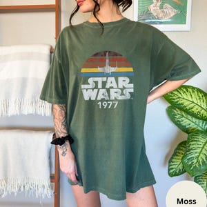 May include: A green oversized t-shirt with a vintage Star Wars graphic. The graphic features a sunset with a spaceship flying through it. The text "Star Wars 1977" is printed below the graphic.