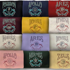 Peut inclure: Une collection de sweatshirts pliés de différentes couleurs, chacun portant le nom et l'emblème d'un dieu grec. Les modèles incluent "Poséidon", "Apollon", "Arès", "Aphrodite", "Dionysos", "Athéna", "Hadès", "Hermès" et "Zeus".