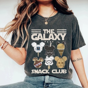 May include: A dark gray t-shirt with a vintage-style graphic featuring the text "The Galaxy Snack Club". The graphic includes cartoon images of popular Star Wars characters, such as Darth Vader, R2-D2, and Yoda, depicted as ice cream pops.