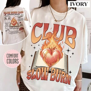 May include: Ivory-colored t-shirt with the text "CLUB SLOW BURN" in orange and red, featuring a heart engulfed in flames above an open book. The back of the shirt has additional graphics and text, including "SLOW BURN CLUB ROMANCE READER".