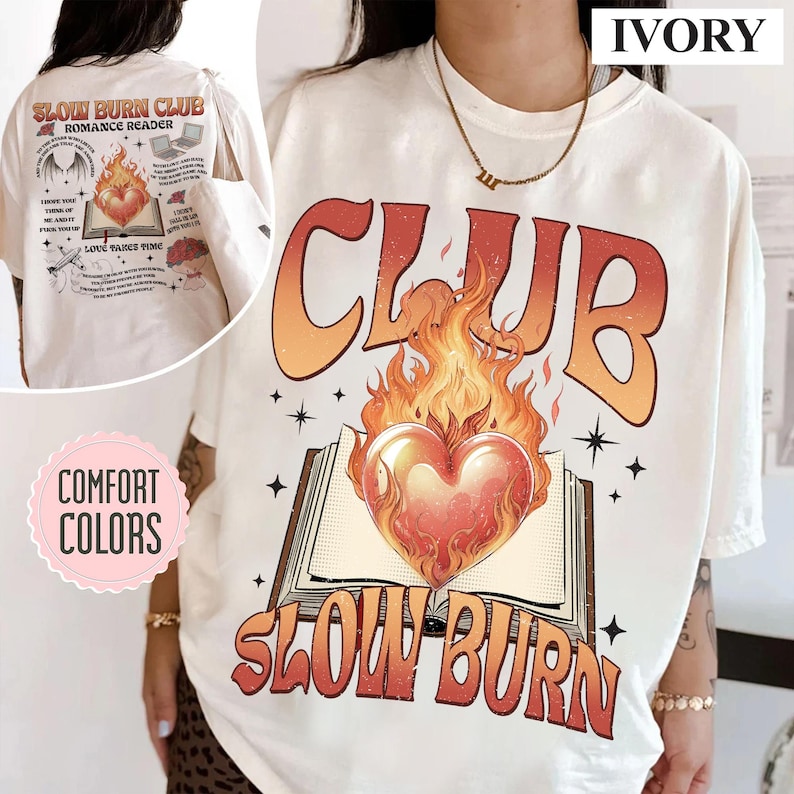 May include: Ivory-colored t-shirt with the text "CLUB SLOW BURN" in orange and red, featuring a heart engulfed in flames above an open book. The back of the shirt has additional graphics and text, including "SLOW BURN CLUB ROMANCE READER".