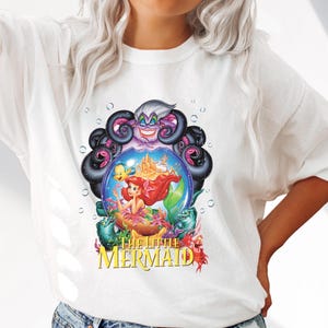 May include: White t-shirt featuring a colorful graphic from Disney's The Little Mermaid. The design includes Ariel, Flounder, Sebastian, Ursula, and a castle. The words "The Little Mermaid" are printed in yellow.