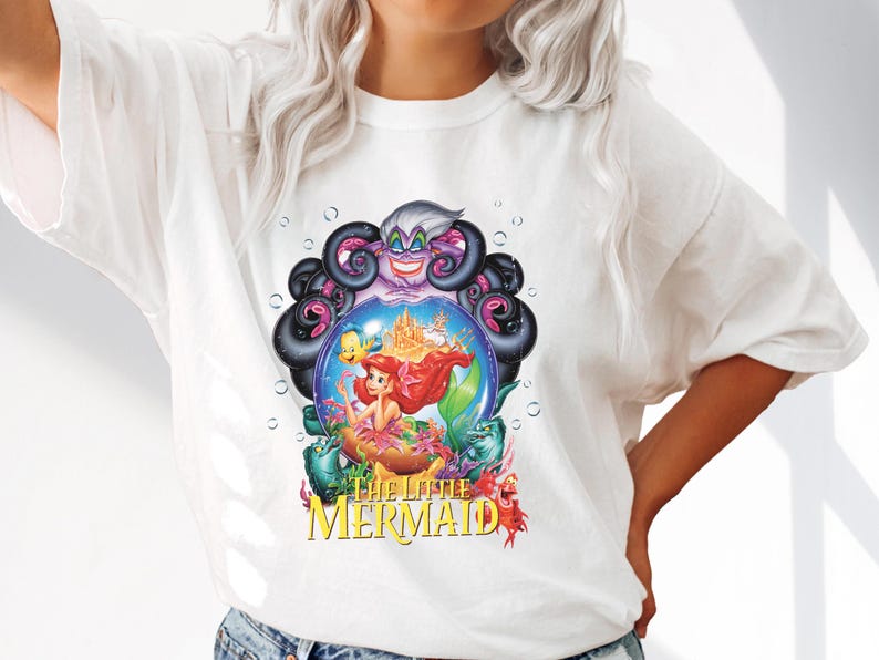 May include: White t-shirt featuring a colorful graphic from Disney's The Little Mermaid. The design includes Ariel, Flounder, Sebastian, Ursula, and a castle. The words "The Little Mermaid" are printed in yellow.