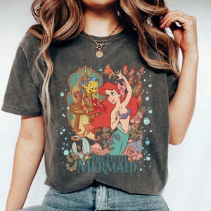 May include: Dark gray t-shirt featuring a colorful graphic of Ariel and Flounder from The Little Mermaid. Ariel has red hair and a purple seashell bra. The shirt has a vintage, faded look.