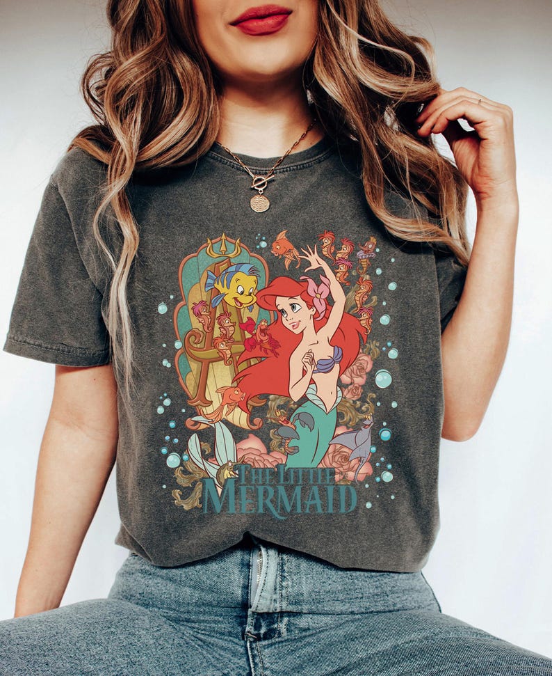May include: Dark gray t-shirt featuring a colorful graphic of Ariel and Flounder from The Little Mermaid. Ariel has red hair and a purple seashell bra. The shirt has a vintage, faded look.