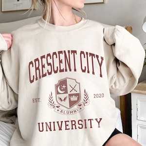 May include: A beige sweatshirt featuring the maroon text "CRESCENT CITY UNIVERSITY". The design includes a crest with a moon, crossed swords, and a crown, with the year 2020. The sweatshirt is a casual style.