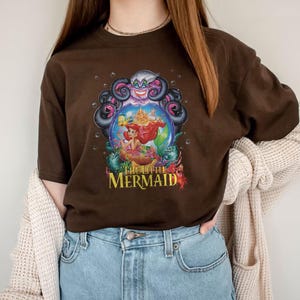 May include: A brown t-shirt featuring a colorful graphic from "The Little Mermaid." The design includes Ariel, Ursula, and other characters from the movie. The text "The Little Mermaid" is in a yellow and red font.
