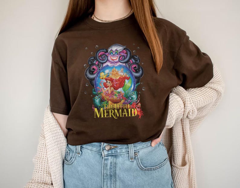May include: A brown t-shirt featuring a colorful graphic from "The Little Mermaid." The design includes Ariel, Ursula, and other characters from the movie. The text "The Little Mermaid" is in a yellow and red font.