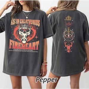 May include: Dark grey t-shirt with a graphic design featuring a deer head, crown, and the text "AN IN CALATHYNIUS FIREHEART". The back of the shirt has a similar design. The shirt is made of a soft material.