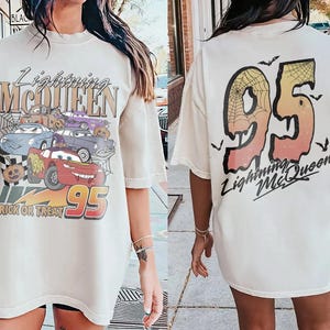 K&ouml;nnte beinhalten: Cremefarbenes Lightning McQueen Grafik-T-Shirt. Die Vorderseite zeigt ein Cartoon-Bild von Lightning McQueen und anderen Autos mit Halloween-Elementen. Auf der R&uuml;ckseite ist eine gro&szlig;e "95" mit Spinnennetz-Design und dem Text "Lightning McQueen" abgebildet.
