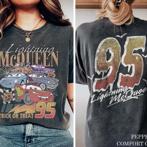 K&ouml;nnte beinhalten: Dunkelgraues T-Shirt mit einem Aufdruck von Lightning McQueen und anderen Cars-Charakteren mit dem Text "Lightning McQueen" und "Trick or Treat 95". Auf der R&uuml;ckseite steht "95 Lightning McQueen" mit Spinnennetz-Design.