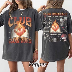 May include: Dark gray t-shirt with a front graphic featuring the text "CLUB SLOW BURN" above an open book with a flaming heart. The back of the shirt has additional text and graphics, including "SLOW BURN CLUB ROMANCE READER".