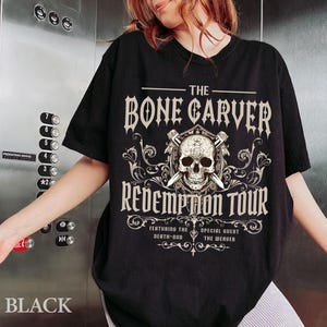 May include: Black t-shirt with a graphic design featuring a skull, crossed swords, and the text "THE BONE CARVER REDEMPTION TOUR". The shirt also includes the text "FEATURING THE DEATH-GOD SPECIAL GUEST THE WEAVER".