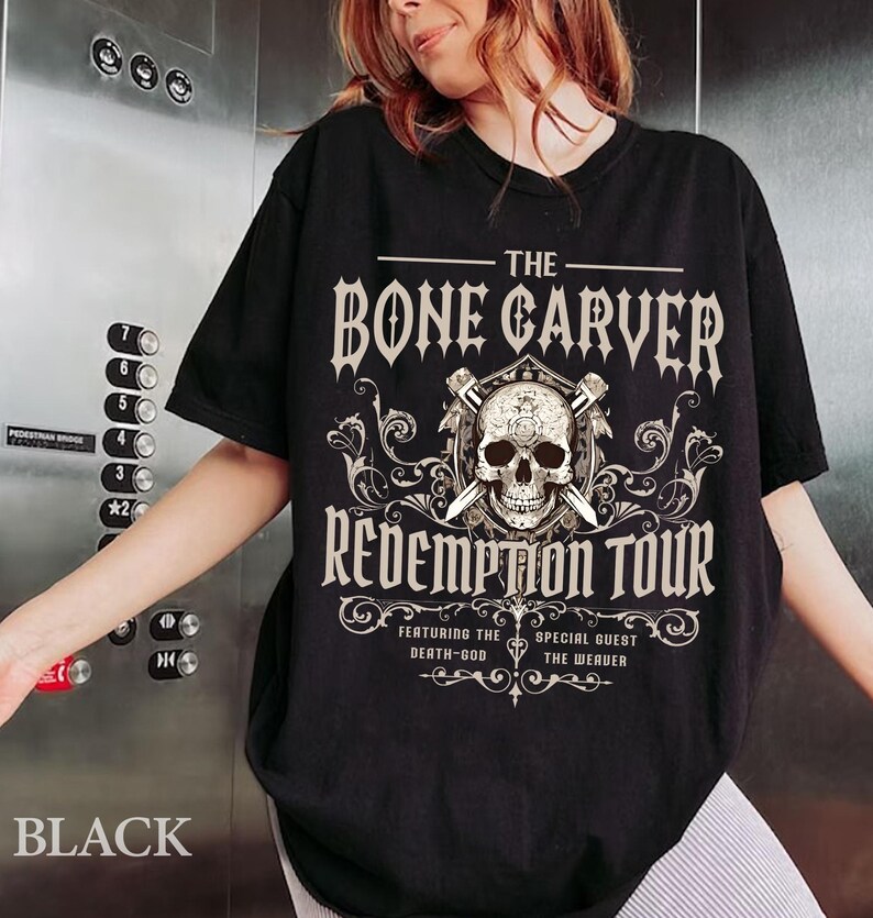 May include: Black t-shirt with a graphic design featuring a skull, crossed swords, and the text "THE BONE CARVER REDEMPTION TOUR". The shirt also includes the text "FEATURING THE DEATH-GOD SPECIAL GUEST THE WEAVER".