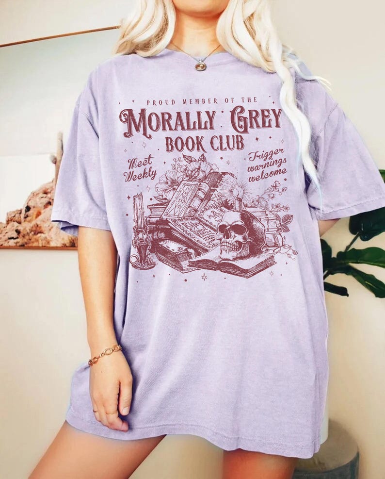 May include: A light purple t-shirt with the text "MORALLY GREY BOOK CLUB" in a burgundy color. The graphic includes a skull, books, and floral elements. The shirt also has the phrases "Meet Weekly" and "Trigger warnings welcome".