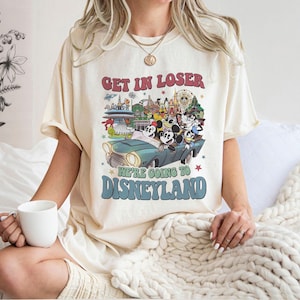 Puede incluir: Camiseta color crema con un gr&aacute;fico retro que dice "GET IN LOSER WE'RE GOING TO DISNEYLAND". El gr&aacute;fico presenta un coche vintage con personajes de Disney y monumentos del parque. La camiseta tiene un corte holgado.