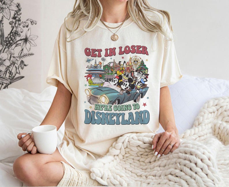 Puede incluir: Camiseta color crema con un gr&aacute;fico retro que dice "GET IN LOSER WE'RE GOING TO DISNEYLAND". El gr&aacute;fico presenta un coche vintage con personajes de Disney y monumentos del parque. La camiseta tiene un corte holgado.
