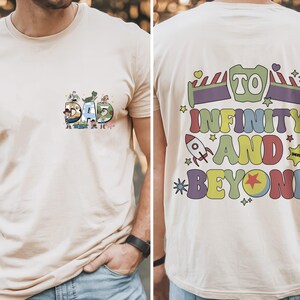 May include: Beige t-shirt with the word "DAD" in colorful letters, each letter featuring cartoon characters. The back of the shirt has the phrase "TO INFINITY AND BEYOND" in a playful, colorful design with a rocket ship and stars.