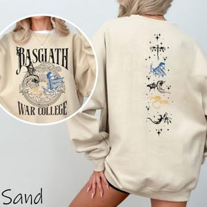 Puede incluir: Sudadera de color arena con un gr&aacute;fico en la parte delantera que dice "Basgiath War College" con ilustraciones de dragones. La parte trasera de la sudadera presenta dise&ntilde;os de dragones en negro, azul y dorado. La sudadera es de color neutro.