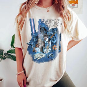 May include: A cream colored t-shirt with a vintage Star Wars graphic print featuring characters like Darth Vader, Luke Skywalker, and Princess Leia. The graphic is in blue, black, and white.
