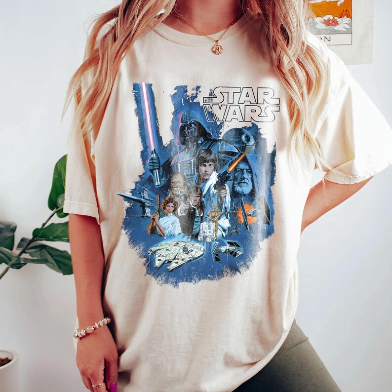 May include: A cream colored t-shirt with a vintage Star Wars graphic print featuring characters like Darth Vader, Luke Skywalker, and Princess Leia. The graphic is in blue, black, and white.
