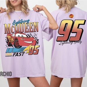 May include: Orchid-colored t-shirt featuring Lightning McQueen from the Cars movie. The front graphic includes the character, the number 95, and the words "Lightning McQueen" and "Think Fast." The back of the shirt displays the number 95 and the words "Lightning Sally."