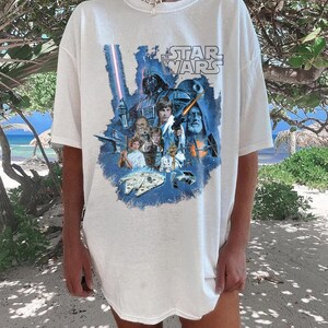 May include: White t-shirt with a blue and white graphic print of characters from the Star Wars movie franchise. The text "Star Wars" is printed in black on the shirt.