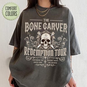 May include: Dark grey t-shirt with the text "THE BONE CARVER REDEMPTION TOUR" and a skull graphic. The shirt also features the text "FEATURING THE DEATH-GOD" and "SPECIAL GUEST THE WEAVER". The shirt has a vintage look and feel.