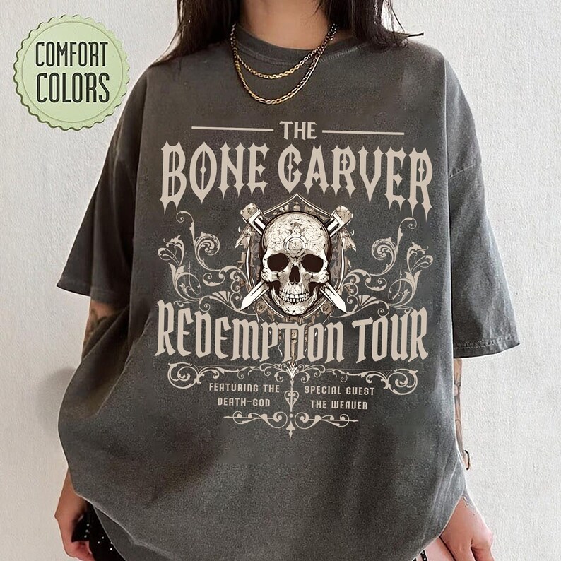May include: Dark grey t-shirt with the text "THE BONE CARVER REDEMPTION TOUR" and a skull graphic. The shirt also features the text "FEATURING THE DEATH-GOD" and "SPECIAL GUEST THE WEAVER". The shirt has a vintage look and feel.