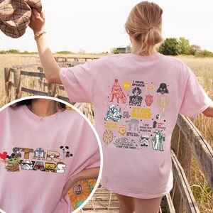 May include: A light pink Star Wars t-shirt with the Star Wars logo and characters like Grogu and R2-D2. The back displays Star Wars graphics and text. The tee shirt is made of a soft material.