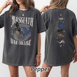 Puede incluir: Camiseta gris oscuro con el texto "BASGIATH WAR COLLEGE" y un gr&aacute;fico de drag&oacute;n. La parte trasera de la camiseta presenta un dise&ntilde;o vertical con m&uacute;ltiples ilustraciones de dragones en varios colores. La palabra "Pepper" est&aacute; impresa en la parte inferior.