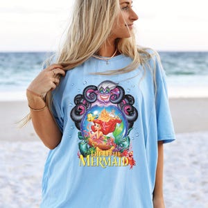May include: Light blue t-shirt featuring a colorful graphic from "The Little Mermaid." The design includes Ariel, Ursula, and other characters. The text "The Little Mermaid" is displayed in yellow. The shirt is worn by a person on a beach.
