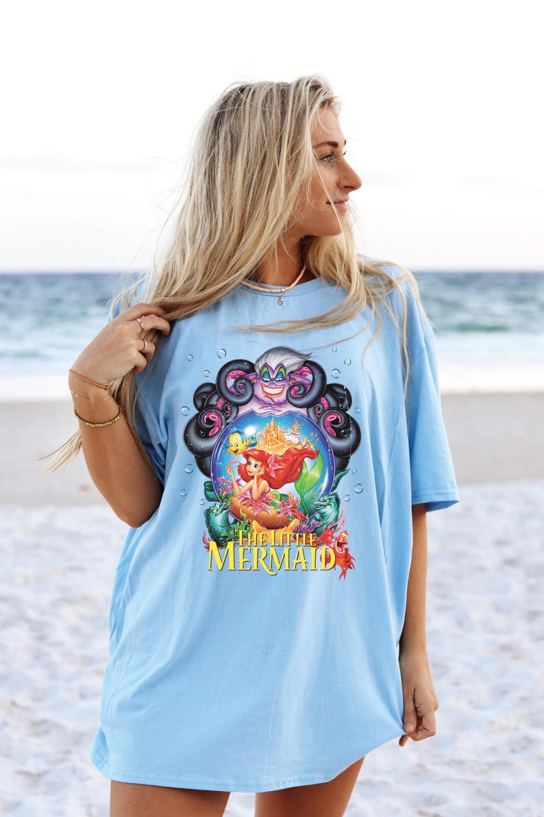 May include: Light blue t-shirt featuring a colorful graphic from "The Little Mermaid." The design includes Ariel, Ursula, and other characters. The text "The Little Mermaid" is displayed in yellow. The shirt is worn by a person on a beach.
