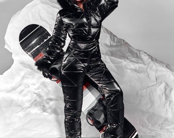 Puede incluir: Una mujer con un traje de esquí negro con capucha forrada de piel y gafas de protección se encuentra de pie con una tabla de snowboard. Lleva guantes negros y botas negras.