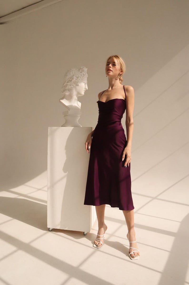 May include: A burgundy midi dress with spaghetti straps. The dress has a cowl neckline and a flowing skirt. The model is wearing white strappy heels. A white bust of a classical figure is on a white pedestal.
