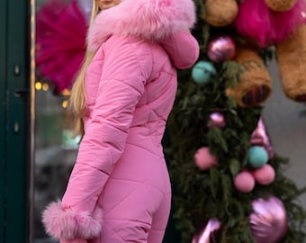 May include: A pink ski suit with a faux fur hood and trim. The suit has a quilted design and is worn by a person standing in front of a decorated Christmas tree.