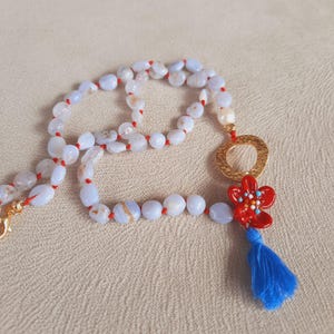 May include: A blue agate stone necklace with a red flower charm and a gold circle pendant. The necklace is strung on red thread and has a blue tassel.