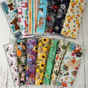 May include: A collection of colorful fabric napkins with various patterns. Designs include cats, sewing machines, stripes, dogs, farm scenes, floral, and bees. The napkins are arranged on a white wooden surface.